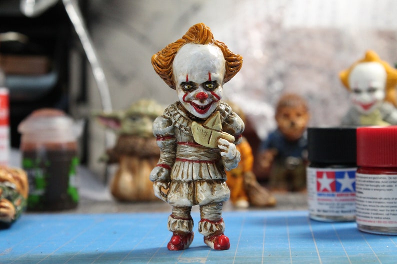 Puede incluir: Una figurita pintada a mano de Pennywise el payaso danzante de la pel&iacute;cula "IT". La figurita lleva un traje blanco y rojo con un barco de papel en la mano. La figurita est&aacute; de pie sobre una superficie azul.
