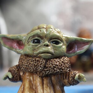 Puede incluir: Una peque&ntilde;a figura verde de un personaje de la franquicia Star Wars, que lleva un abrigo de piel marr&oacute;n. La figura tiene orejas grandes y ojos grandes.