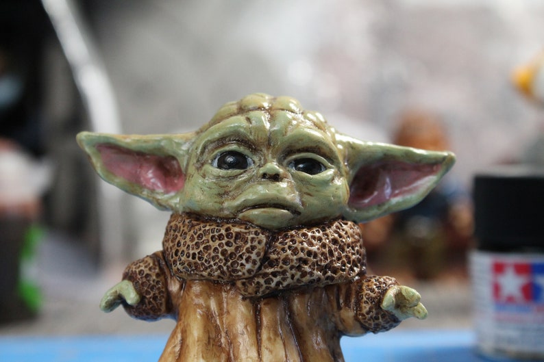 Puede incluir: Una peque&ntilde;a figura verde de un personaje de la franquicia Star Wars, que lleva un abrigo de piel marr&oacute;n. La figura tiene orejas grandes y ojos grandes.