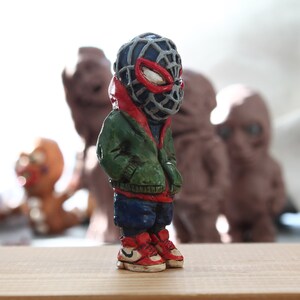Puede incluir: Una peque&ntilde;a figurita pintada a mano de un superh&eacute;roe que lleva una m&aacute;scara roja y negra, una chaqueta verde, pantalones azules y zapatillas rojas y blancas. La figurita est&aacute; de pie sobre una superficie de madera.