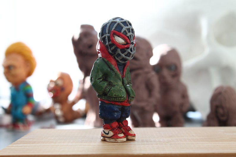 Puede incluir: Una peque&ntilde;a figurita pintada a mano de un superh&eacute;roe que lleva una m&aacute;scara roja y negra, una chaqueta verde, pantalones azules y zapatillas rojas y blancas. La figurita est&aacute; de pie sobre una superficie de madera.