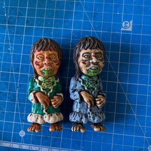 May include: Two small, hand-painted figurines of children with green and blue pajamas. The figurines have green and white painted faces with exaggerated features and are wearing green and blue pajamas with white trim.