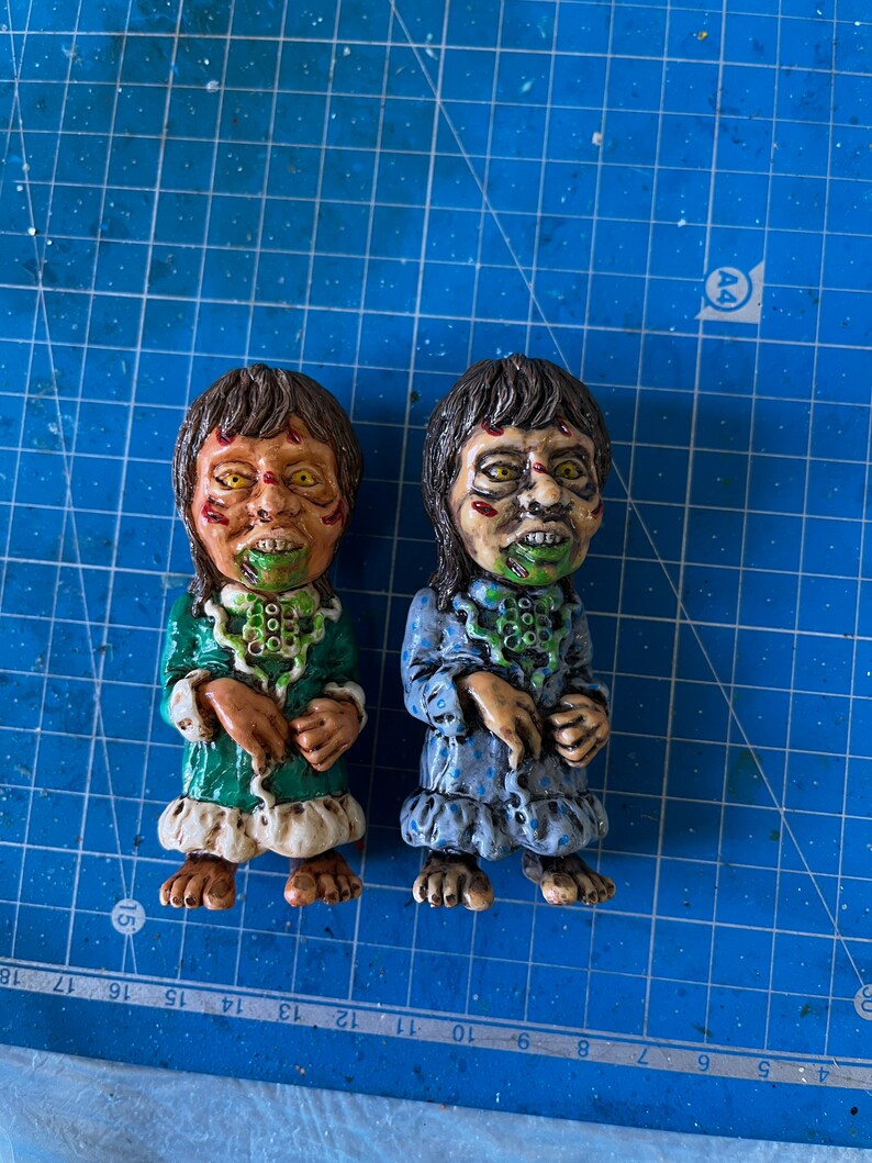 May include: Two small, hand-painted figurines of children with green and blue pajamas. The figurines have green and white painted faces with exaggerated features and are wearing green and blue pajamas with white trim.