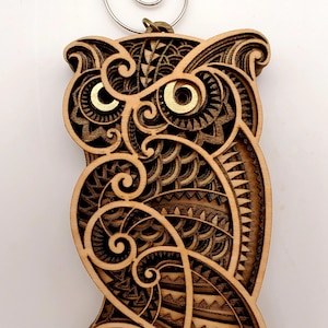 May include: A wooden owl ornament with intricate laser-cut details. The owl is brown with gold accents around the eyes.