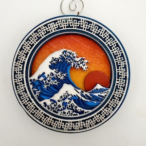 May include: A round wooden ornament with a blue and white wave design and a red sun. The ornament is surrounded by a decorative border.