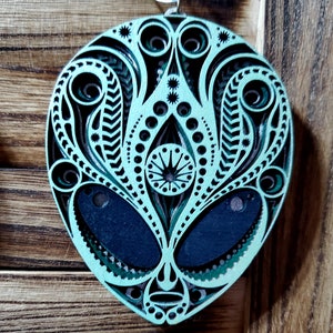 May include: A teal green alien face ornament with intricate black and green patterns. The ornament is made of wood and has a silver hanger.