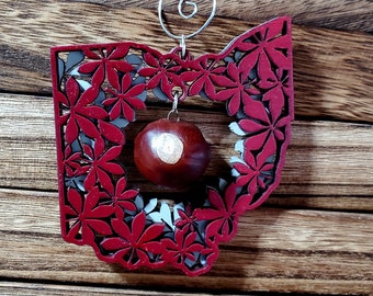 Op de afbeelding: Rood houten ornament in de vorm van de staat Ohio met een uitgesneden ontwerp van bladeren. Een bruine kastanje hangt in het midden.