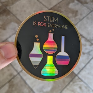 May include: A round sticker with a black background and gold trim. The sticker says "STEM is for everyone" in gold lettering. There are four colorful beakers with rainbow stripes on the sticker.