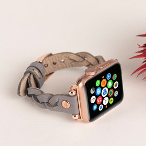 May include: Rose gold Apple Watch with a gray braided leather band. The watch face displays colorful app icons. The band features rose gold hardware and a braided design, adding a stylish touch to the wearable technology.