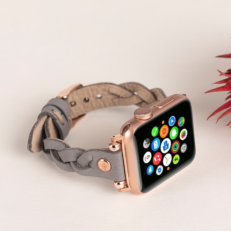 May include: Rose gold Apple Watch with a gray braided leather band. The watch face displays colorful app icons. The band features rose gold hardware and a braided design, adding a stylish touch to the wearable technology.