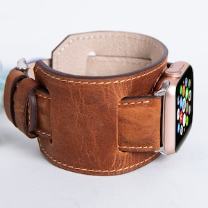 Könnte beinhalten: Ein braunes Lederarmband für eine Apple Watch. Das Armband ist aus echtem Leder gefertigt und hat einen genähten Rand. Das Armband ist so konzipiert, dass es um das Handgelenk passt und mit einer Schnalle geschlossen wird.