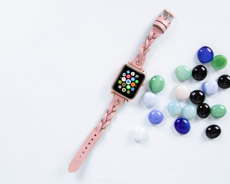 K&ouml;nnte beinhalten: Ein rosafarbenes geflochtenes Lederarmband f&uuml;r eine Apple Watch mit ros&eacute;goldener Oberfl&auml;che. Das Zifferblatt der Uhr ist sichtbar und zeigt den Apple Watch-Startbildschirm mit verschiedenen App-Symbolen.
