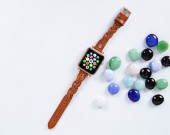May include: A brown leather braided watch strap with a rose gold Apple Watch displaying the home screen. The watch is on a light blue background with scattered blue, green and white glass stones.