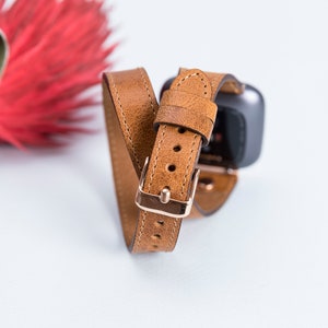 May include: A brown leather watch band with a rose gold buckle. The band is designed to wrap around the wrist twice.