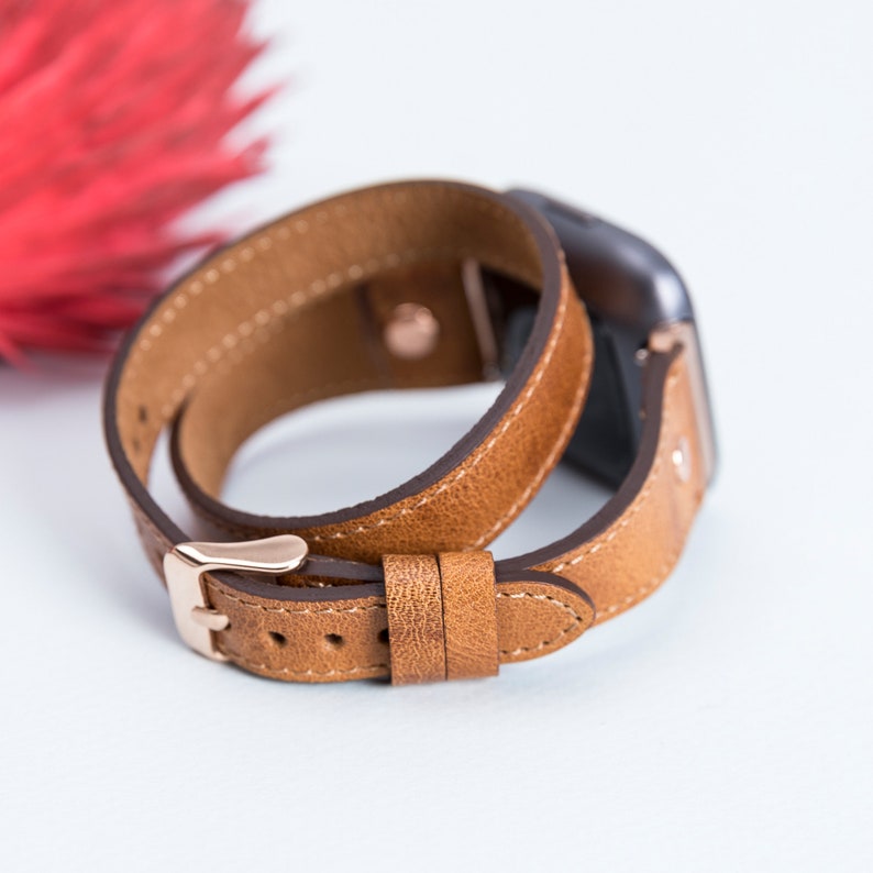May include: A brown leather watch band with a rose gold buckle. The band is double-wrapped and has a textured finish.