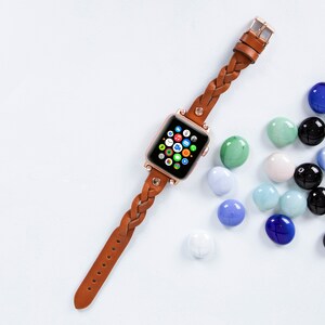 May include: A brown leather braided watch band with a rose gold Apple Watch with the home screen displayed. The watch is on a light blue background with scattered blue, green, and white glass stones.
