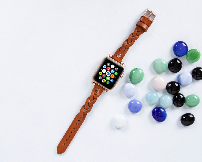 May include: A brown leather braided watch band with a rose gold Apple Watch with the home screen displayed. The watch is on a light blue background with scattered blue, green, and white glass stones.