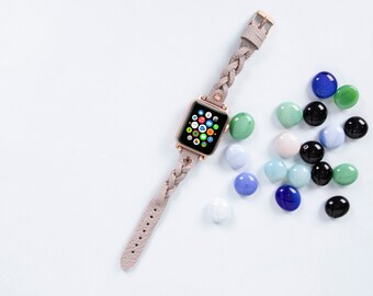 May include: A rose gold Apple Watch with a light grey braided leather strap. The watch face is on and displays a grid of colourful app icons. The watch is surrounded by small, colourful glass stones.