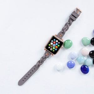 May include: A rose gold smartwatch with a gray braided leather band. The watch face displays colorful app icons. The band has a buckle closure. Colorful glass marbles in shades of blue, green, and black are scattered around the watch.