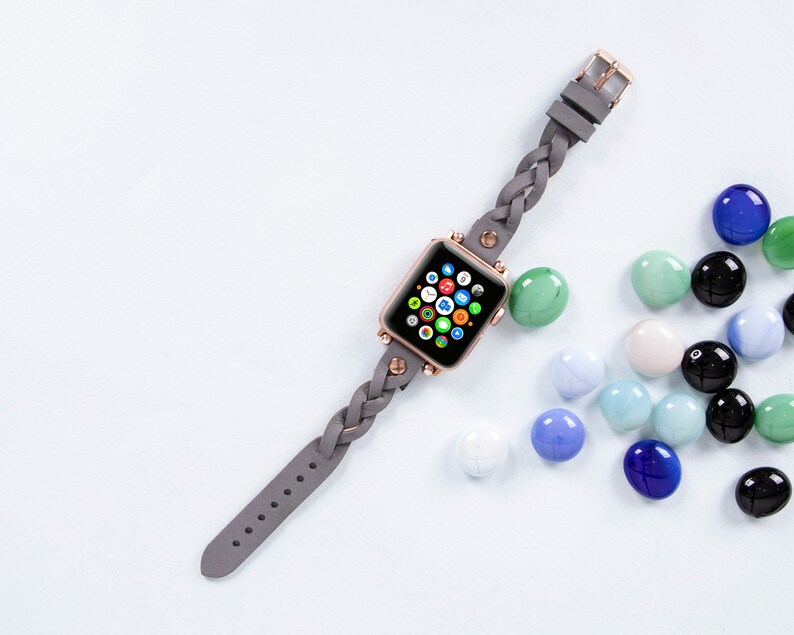 May include: A rose gold smartwatch with a gray braided leather band. The watch face displays colorful app icons. The band has a buckle closure. Colorful glass marbles in shades of blue, green, and black are scattered around the watch.