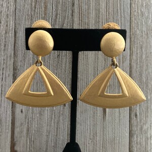 May include: A pair of gold tone clip-on earrings with a geometric triangle design. The earrings have a smooth, matte finish.