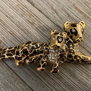 May include: A gold-tone brooch featuring two leopards, one larger than the other, with black and brown spots. The leopards are adorned with clear rhinestones.