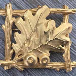 May include: A gold-toned brooch featuring an oak leaf and two acorns within a rectangular frame made of twig-like elements. The brooch has a detailed, textured surface, and the overall design evokes a natural, rustic aesthetic.