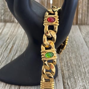 May include: A gold-toned chain bracelet with red and green oval-shaped gemstones. The bracelet features a chunky curb chain design with rectangular accents. The bracelet is displayed on a black hand-shaped jewelry holder.