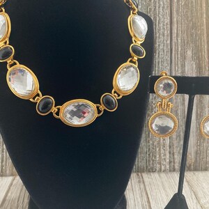 Pode incluir: Um conjunto de colar e brincos em tom dourado com grandes pedras ovais transparentes e pretas.
