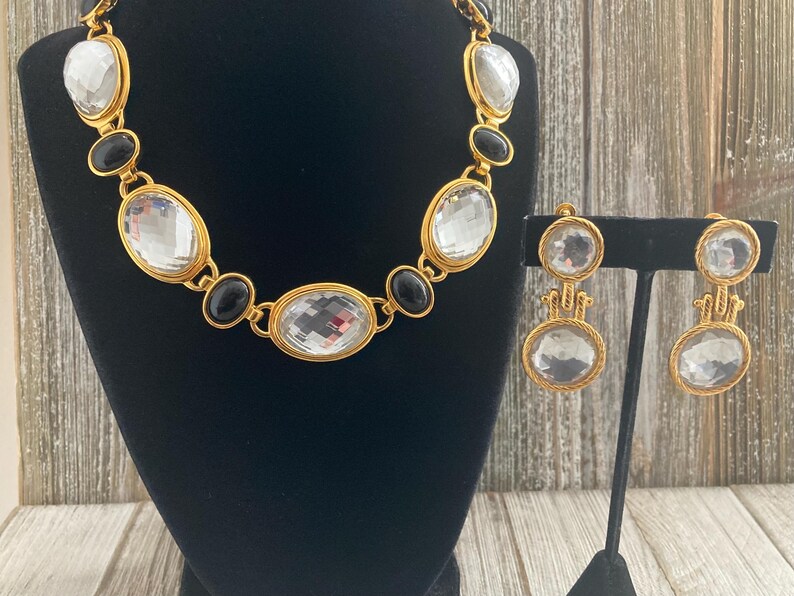 Pode incluir: Um conjunto de colar e brincos em tom dourado com grandes pedras ovais transparentes e pretas.