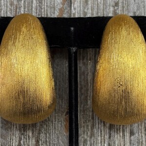 May include: A pair of gold-colored clip-on earrings. The earrings have a textured surface and a teardrop shape. The earrings are displayed on a black velvet earring holder against a wooden background.