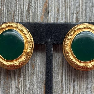 K&ouml;nnte beinhalten: Ein Paar goldfarbene Clip-Ohrringe mit einem dunkelgr&uuml;nen Mittelteil. Die Ohrringe haben eine runde Form mit einem dekorativen, strukturierten Rand. Die Ohrringe werden auf einem schwarzen St&auml;nder vor einem Holzgrund pr&auml;sentiert.