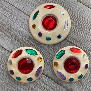 May include: Three white ceramic buttons with a raised center and a red jewel in the center. The buttons are decorated with colorful jewels around the center jewel.