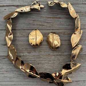 Pu&ograve; includere: Un set di gioielli color oro composto da una collana e orecchini abbinati. La collana &egrave; composta da elementi a forma di foglia collegati. Gli orecchini sono ovali con una superficie strutturata. Il set &egrave; esposto su una superficie di legno.