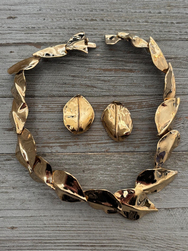 Pu&ograve; includere: Un set di gioielli color oro composto da una collana e orecchini abbinati. La collana &egrave; composta da elementi a forma di foglia collegati. Gli orecchini sono ovali con una superficie strutturata. Il set &egrave; esposto su una superficie di legno.