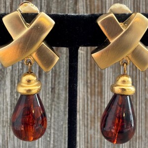 May include: Gold-tone clip-on earrings with a cross design and teardrop-shaped amber-colored pendants. The earrings are displayed on a black velvet stand against a wooden background. The jewelry has a vintage aesthetic.