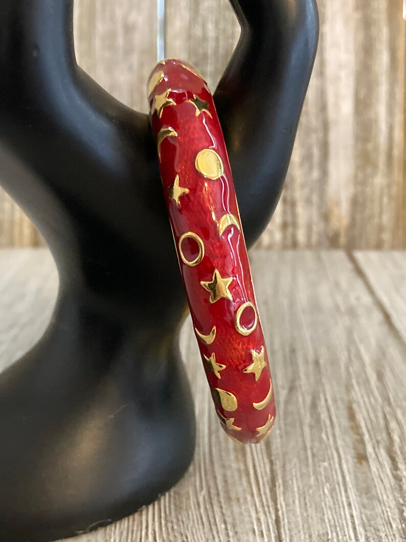 May include: Red enamel bangle bracelet with a gold design of stars, moons, and circles.