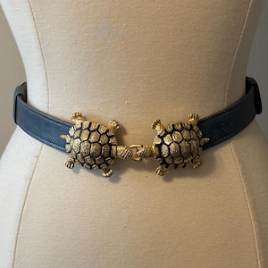 Op de afbeelding: Een donkerblauwe riem met textuur en een decoratieve goudkleurige metalen gesp in de vorm van twee in elkaar grijpende schildpadden. De schildpadden hebben gedetailleerde schildpatronen en zijn in het midden verbonden als sluiting.