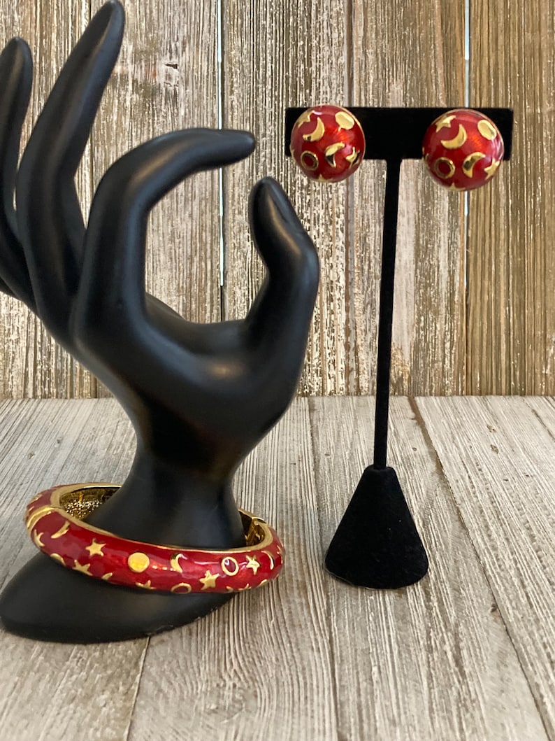 May include: A red and gold bangle bracelet and matching earrings with a crescent moon and star pattern. The jewelry is displayed on a black mannequin hand and stand.