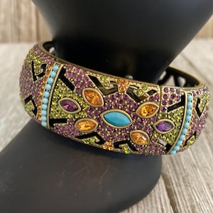 Könnte beinhalten: Ein goldfarbenes Armreif-Armband mit geometrischem Design. Das Armband ist mit violetten, grünen, orangefarbenen und blauen Strasssteinen verziert.