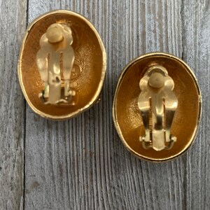 May include: A pair of gold-tone oval-shaped clip-on earrings. The earrings have a textured surface and a simple design.