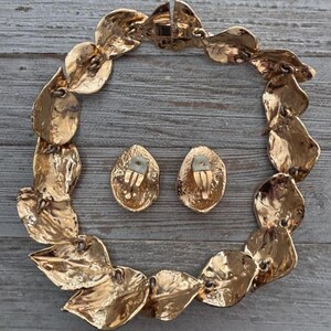 Pu&ograve; includere: Un set di collana e orecchini a clip abbinati, color oro. La collana &egrave; composta da maglie a forma di foglia e gli orecchini sono ovali. Il set di gioielli &egrave; esposto su una superficie di legno grigio e invecchiato.