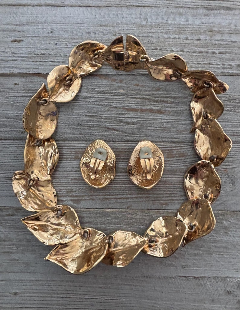 Pu&ograve; includere: Un set di collana e orecchini a clip abbinati, color oro. La collana &egrave; composta da maglie a forma di foglia e gli orecchini sono ovali. Il set di gioielli &egrave; esposto su una superficie di legno grigio e invecchiato.