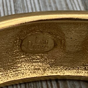May include: Close-up of the inside of a gold-toned bangle bracelet. The bracelet is stamped with the words "Joan Rivers" in a circular oval.