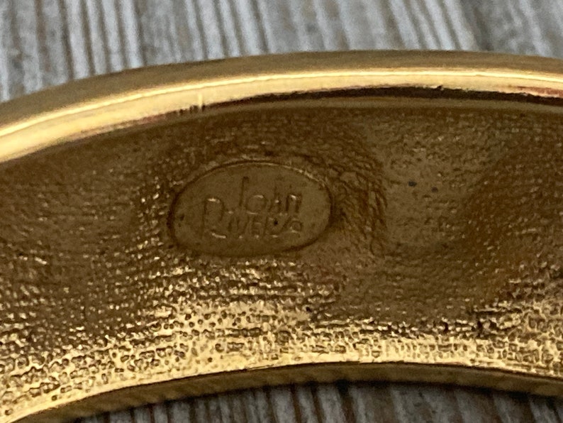 May include: Close-up of the inside of a gold-toned bangle bracelet. The bracelet is stamped with the words "Joan Rivers" in a circular oval.