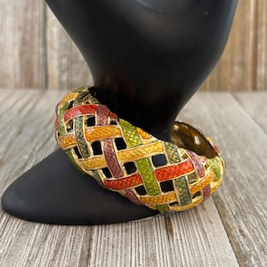 May include: A colorful enamel bracelet with a woven design. The bracelet features a mix of red, green, yellow, and purple hues. The gold-toned metal bracelet is displayed on a black stand against a wooden background.