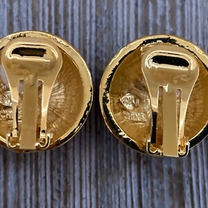 May include: A pair of gold-tone clip-on earrings with a round design. The back of the earrings have a gold-tone clasp and the words "Made in China" are engraved on the back.