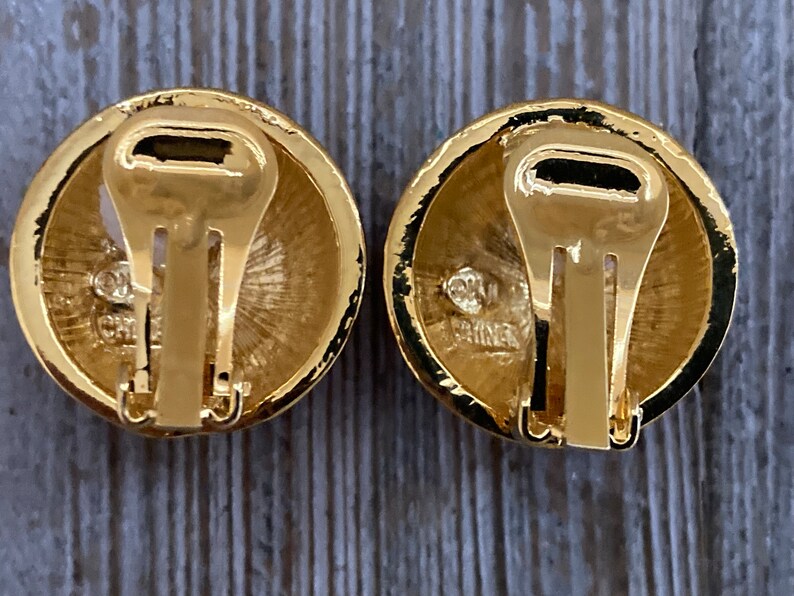 May include: A pair of gold-tone clip-on earrings with a round design. The back of the earrings have a gold-tone clasp and the words "Made in China" are engraved on the back.