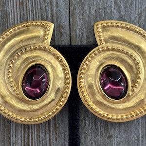 Puede incluir: Un par de pendientes dorados con un diseño en espiral y una piedra central ovalada de color púrpura oscuro. Los pendientes tienen un borde con cuentas y un acabado pulido, adecuados para ocasiones formales.