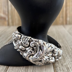 Puede incluir: Una pulsera plateada con bisagras y un diseño floral ornamentado, que incluye rosas y hojas. La pulsera se muestra en un brazo de maniquí negro sobre un fondo de madera. La pulsera tiene una cadena.